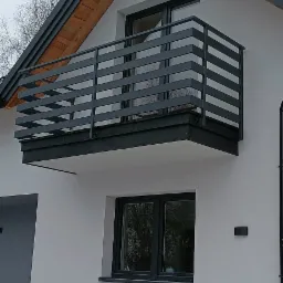 Nowoczesny balkon z metalową balustradą o poziomym układzie szczebli, przymocowany do ściany domu o jasnej elewacji, widok z dołu na tle pochmurnego nieba.