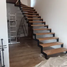 Nowoczesne schody drewniane z czarną metalową konstrukcją w trakcie montażu, zabezpieczone folią malarską, widoczne drabiny w tle, Mirów Stary.