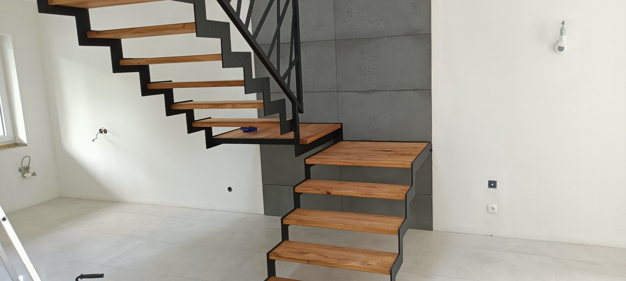 Nowoczesne schody z drewnianymi stopniami i czarną, metalową konstrukcją, na tle szarej ściany z betonowych płyt, widoczne niedokończone instalacje elektryczne.