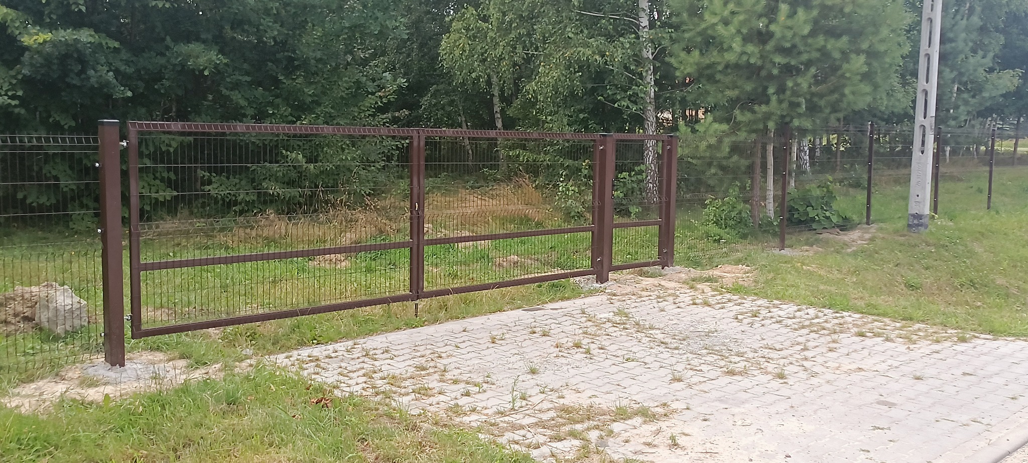 Brązowa, dwuskrzydłowa brama z siatki panelowej na posesji z betonową kostką, widok na las w tle.