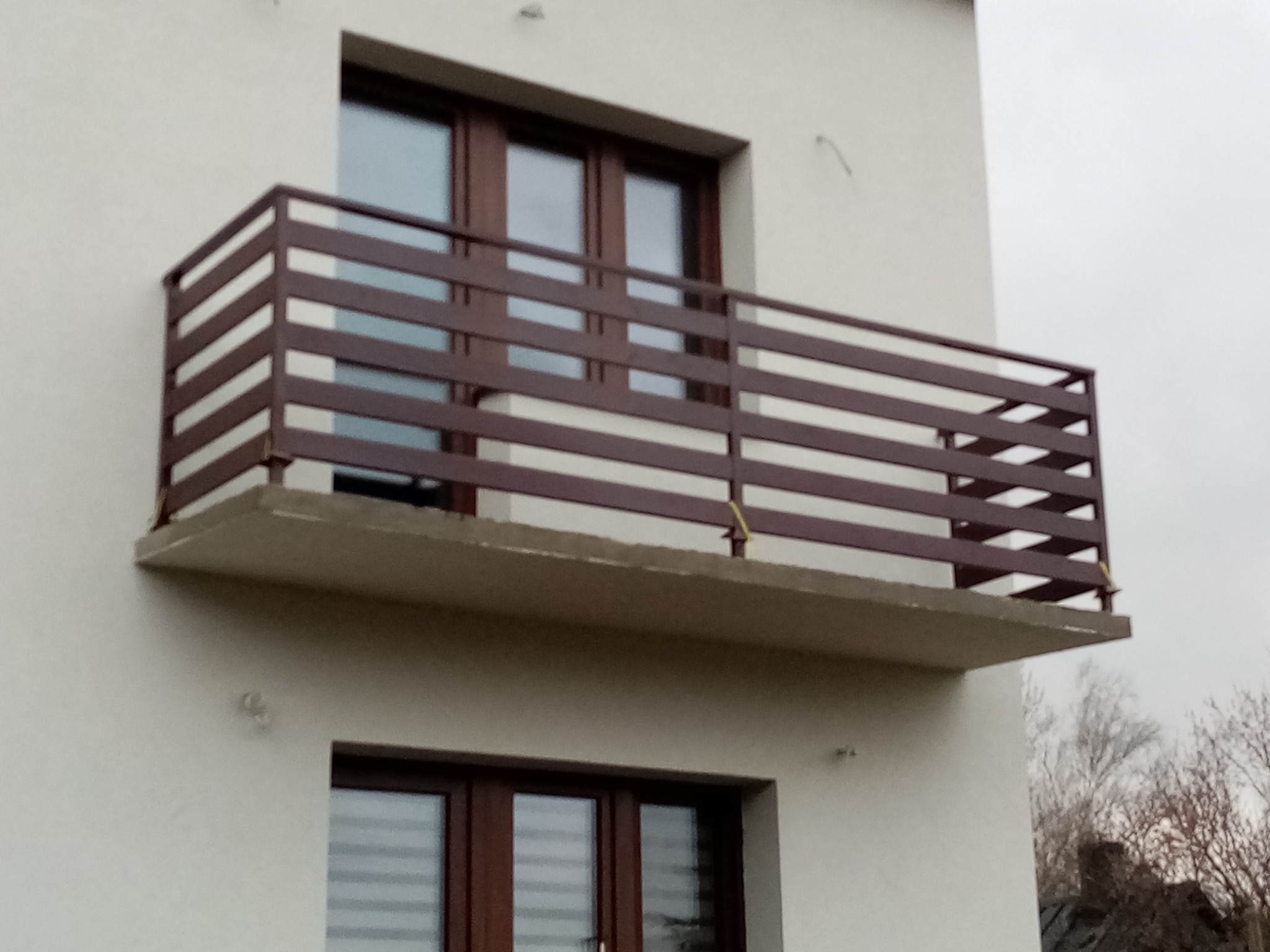 Balkon z brązową metalową balustradą z poziomych profili, widok z dołu na tle białej elewacji i pochmurnego nieba.
