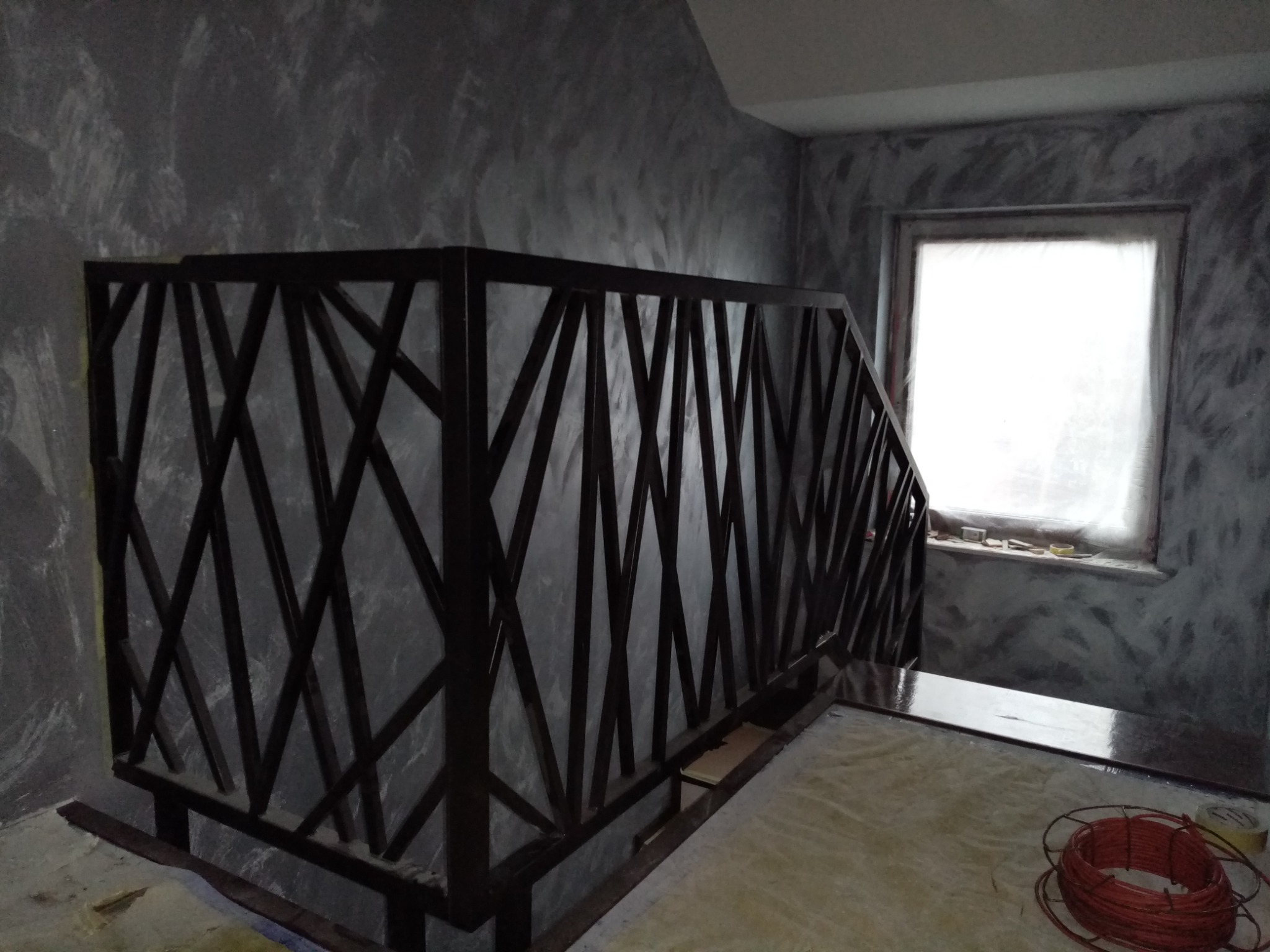 Nowoczesna, metalowa balustrada schodowa z geometrycznym wzorem krzyżujących się prętów, na tle ściany o fakturze betonu architektonicznego i okna z widocznym parapetem.