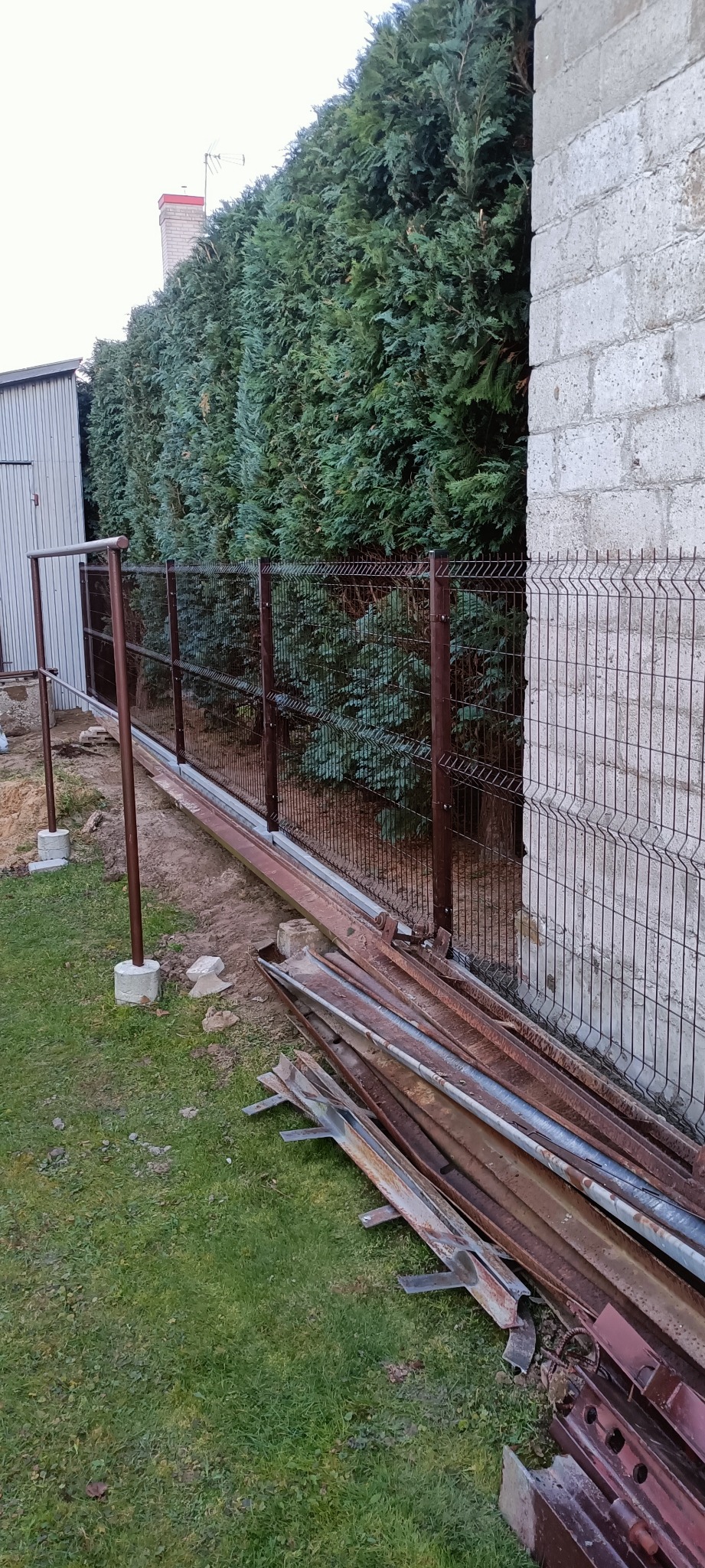Montaż brązowego ogrodzenia z paneli siatkowych na betonowej podmurówce, widoczne słupki i elementy montażowe, w tle wysokie tuje i budynek z bloczków betonowych.