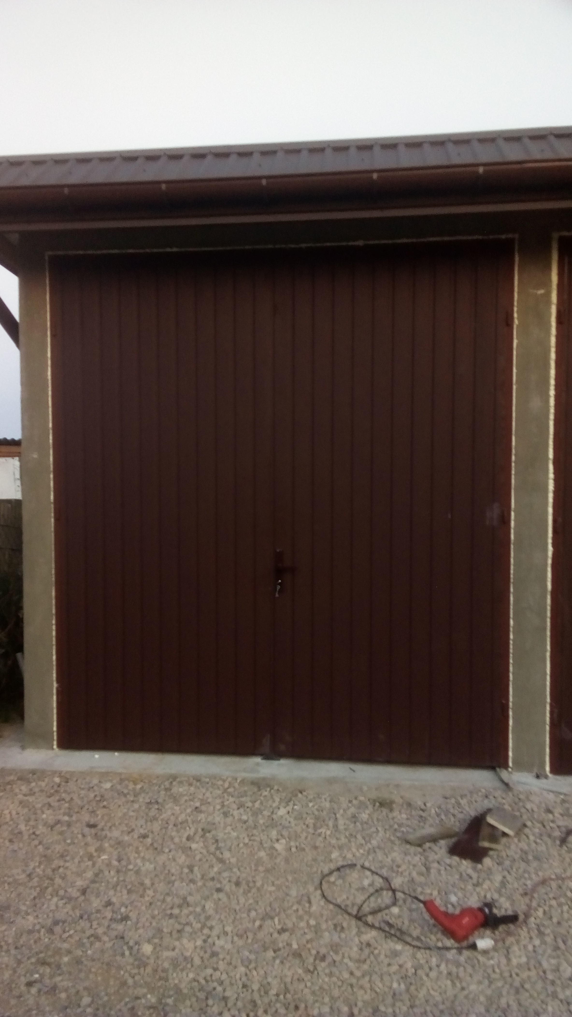 Brązowa brama garażowa z pionowymi panelami, widziana z zewnątrz na tle żwirowego podjazdu, z widocznym narzędziem elektrycznym leżącym obok.