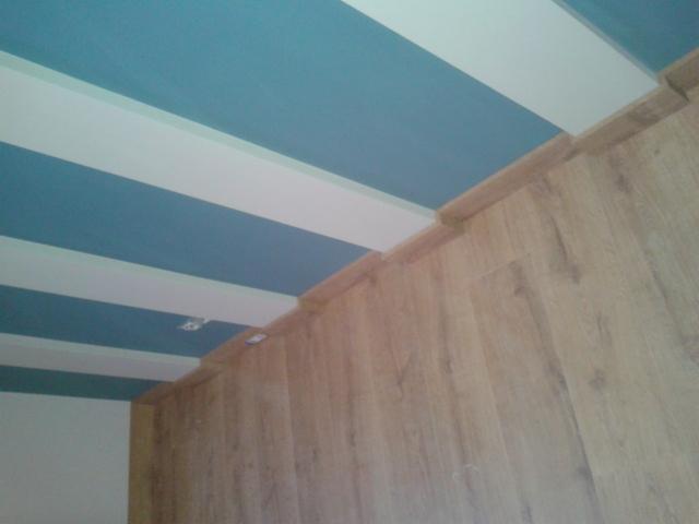 Ściana wyłożona panelami podłogowymi z widocznym fragmentem sufitu w biało-niebieskie pasy, listwa przypodłogowa wykończona w kolorze paneli.