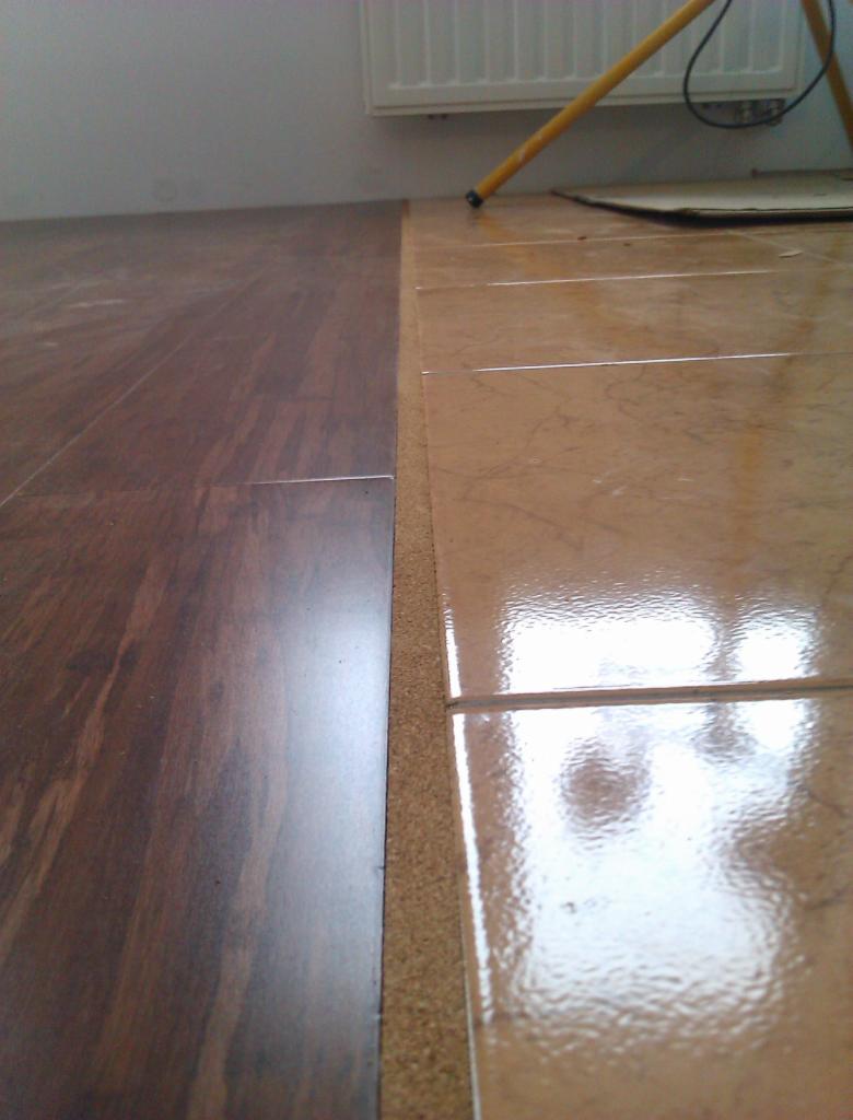 Połączenie ciemnych paneli podłogowych i jasnych płytek ceramicznych za pomocą korkowej listwy dylatacyjnej, widoczny fragment radiatora i lampy roboczej.