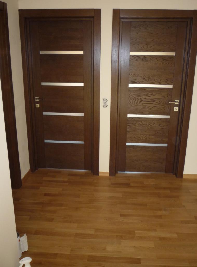 Dwa brązowe, nowoczesne skrzydła drzwiowe z poziomymi, srebrnymi intarsjami, zamontowane w jasnym wnętrzu z drewnianą podłogą.