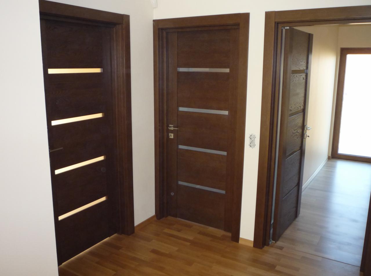 Trzy brązowe drzwi wewnętrzne z poziomymi przeszkleniami, zamontowane w korytarzu z drewnianą podłogą.