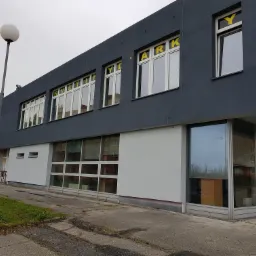 Dwukondygnacyjny budynek z szarą elewacją i białym parterem, z żółtymi literami na oknach tworzącymi napisy, widoczna lampa uliczna i fragment trawnika.