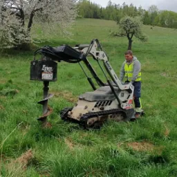 Operator w kamizelce odblaskowej obsługuje wiertnicę Bobcat MT50 wiercącą otw&oacute;r w trawiastym terenie pod przyszłe ogrodzenie, w tle kwitnące drzewo i zalesione wzg&oacute;rze.