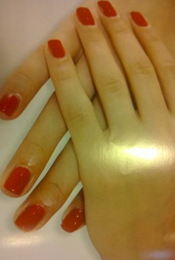 Dłonie z czerwonym, kwadratowym manicure, jedna dłoń ułożona na drugiej, widoczne delikatne refleksy światła na skórze.