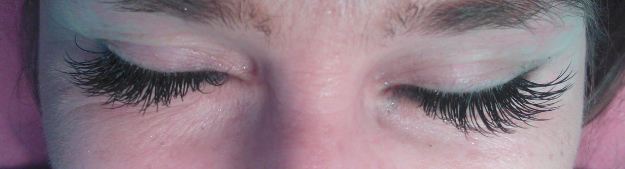 Zbliżenie na zamknięte oczy z długimi, czarnymi, sztucznymi rzęsami, z widocznymi drobnymi niedoskonałościami skóry wokół oczu i brwi.