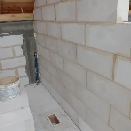 Wznoszenie ściany z białych bloczków, widoczny poziomica, wiadro z zaprawą i drewniany szkielet w podłodze.