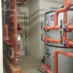 Kompleksowa instalacja grzewcza w kotłowni: czerwone rury, zawory z niebieskimi akcentami, zbiornik akumulacyjny z logo, widoczne elementy automatyki i czujniki.