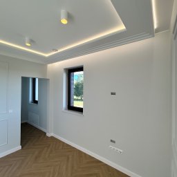 MICH-REM - Jasne wnętrze po remoncie: parkiet w jodełkę, białe ściany z gniazdkami, okno z widokiem na zieleń i podwieszany sufit z oświetleniem LED.