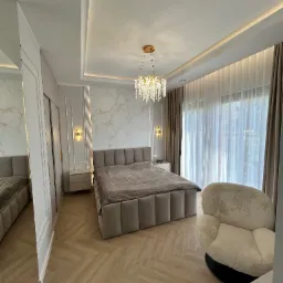 Elegancka sypialnia z łóżkiem kontynentalnym, parkietem w jodełkę i kryształowym żyrandolem. Marmurowa ściana za wezgłowiem, beżowe zasłony i fotel dodają przytulności.