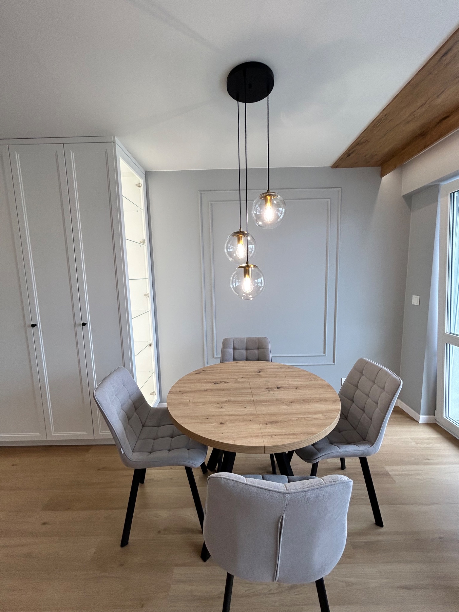 Nowoczesna aranżacja jadalni: okrągły stół z drewnianym blatem, szare krzesła z pikowaniem, wisząca lampa z trzema szklanymi kloszami, zabudowa meblowa w odcieniach szarości.
