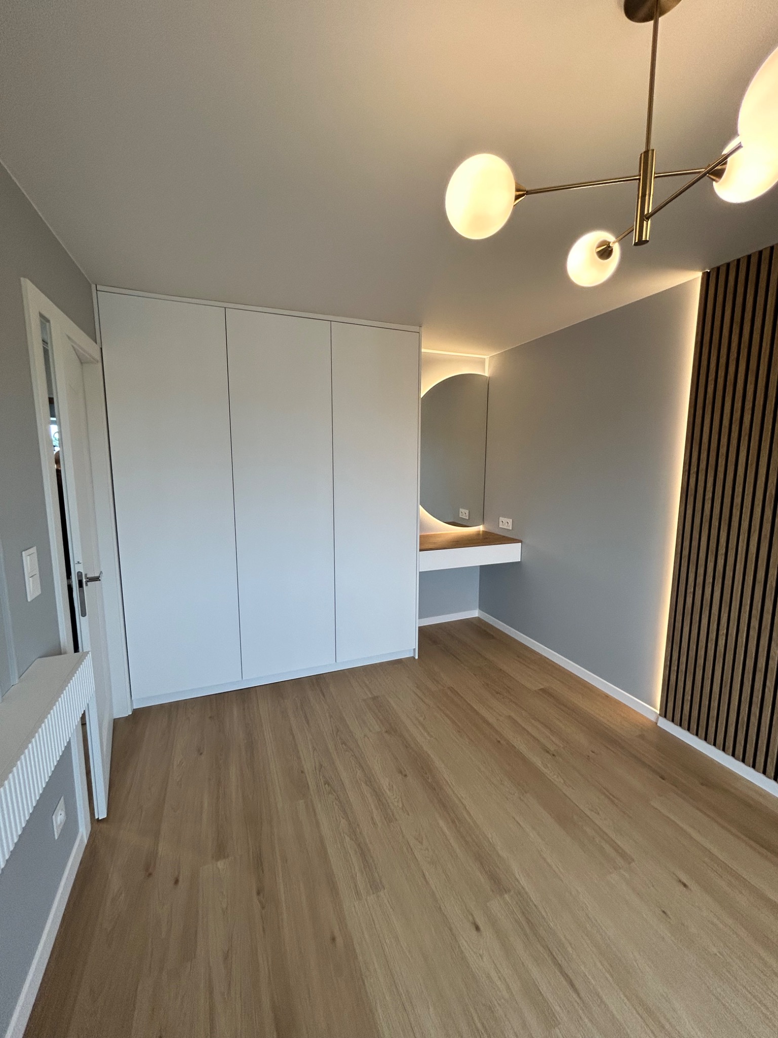 Nowoczesna sypialnia po remoncie: biała szafa wnękowa, drewniana podłoga, toaletka z okrągłym lustrem podświetlonym LED, dekoracyjne panele ścienne.