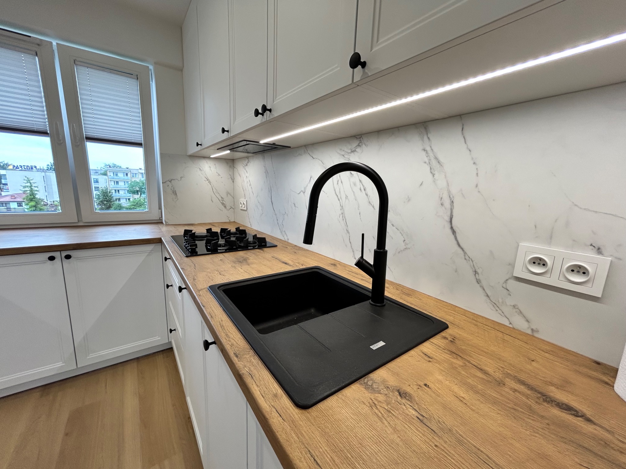 Nowoczesna kuchnia z białymi szafkami, drewnianym blatem i czarnym zlewem na tle marmurowej ściany. Nad blatem oświetlenie LED.