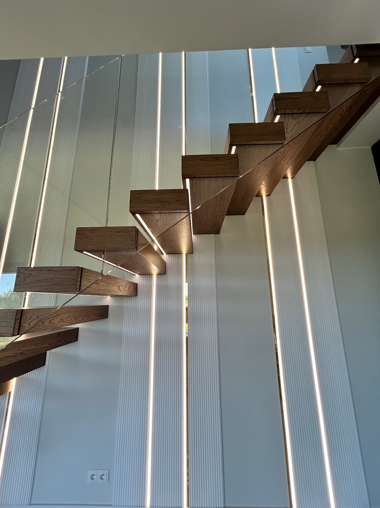 Nowoczesne, drewniane schody wspornikowe z podświetleniem LED na białej ścianie z dekoracyjnymi panelami. Minimalistyczny design.