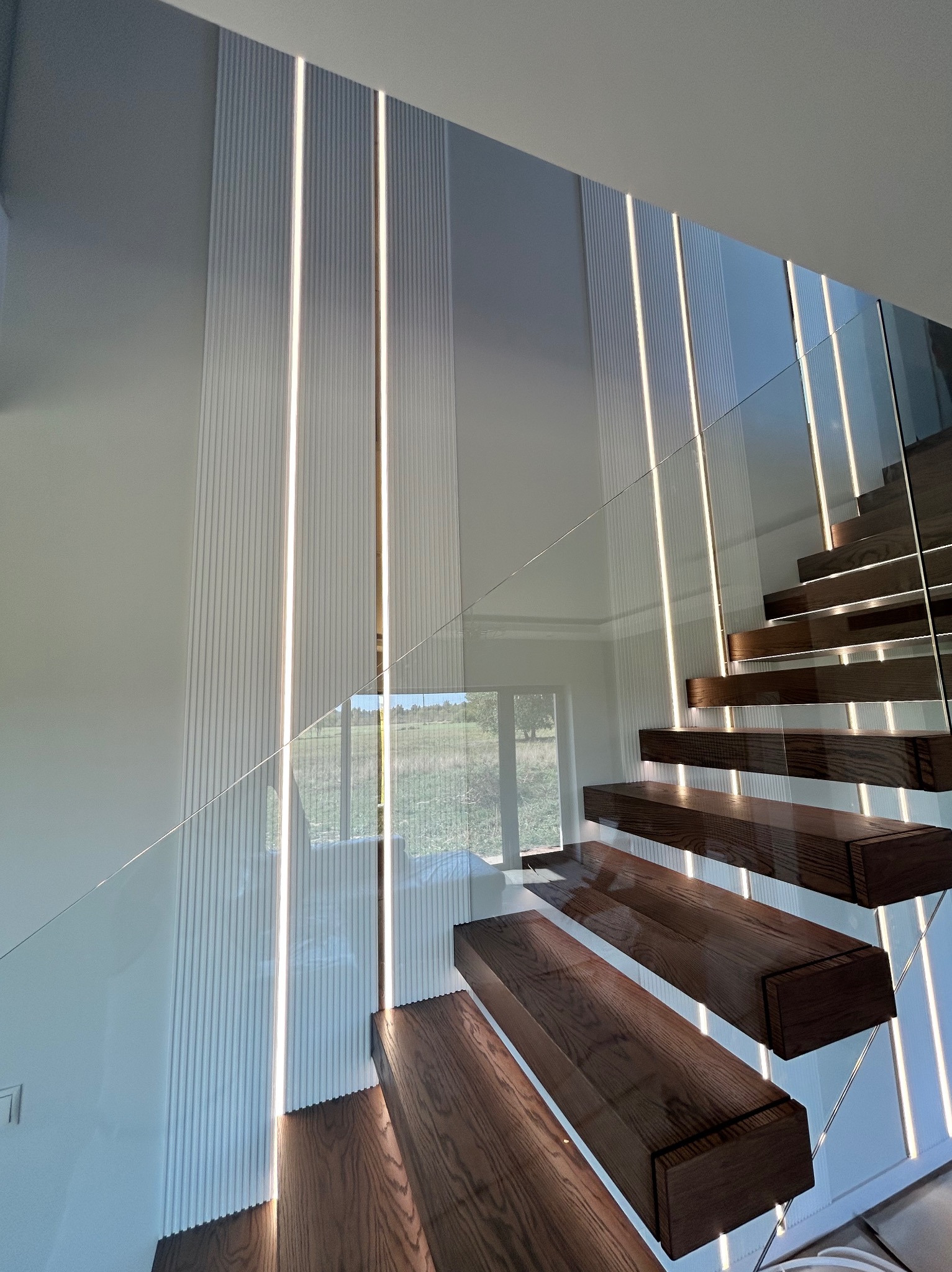 Nowoczesne schody z drewnianymi stopniami i szklaną balustradą, podświetlone pionowymi listwami LED na ścianie. Widok z dołu.