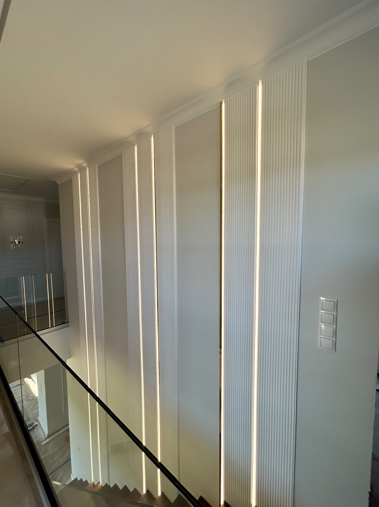 Nowoczesna ściana z dekoracyjnymi panelami i subtelnym oświetleniem LED, widok z góry na schody z szklaną balustradą. Eleganckie wykończenie wnętrza.