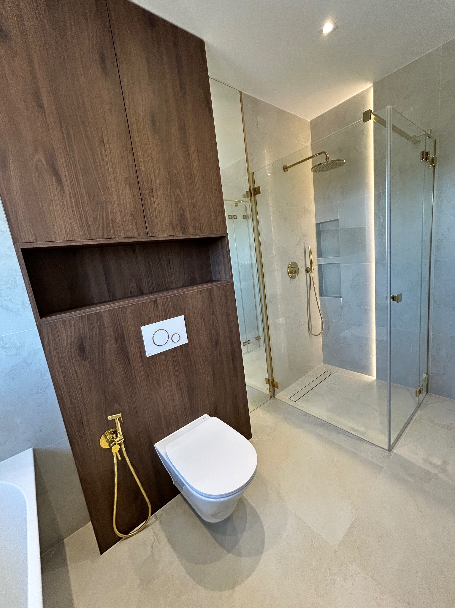 Nowoczesna łazienka z kabiną prysznicową ze złotymi detalami, drewnianą zabudową WC i bidetką. Stonowana kolorystyka płytek.