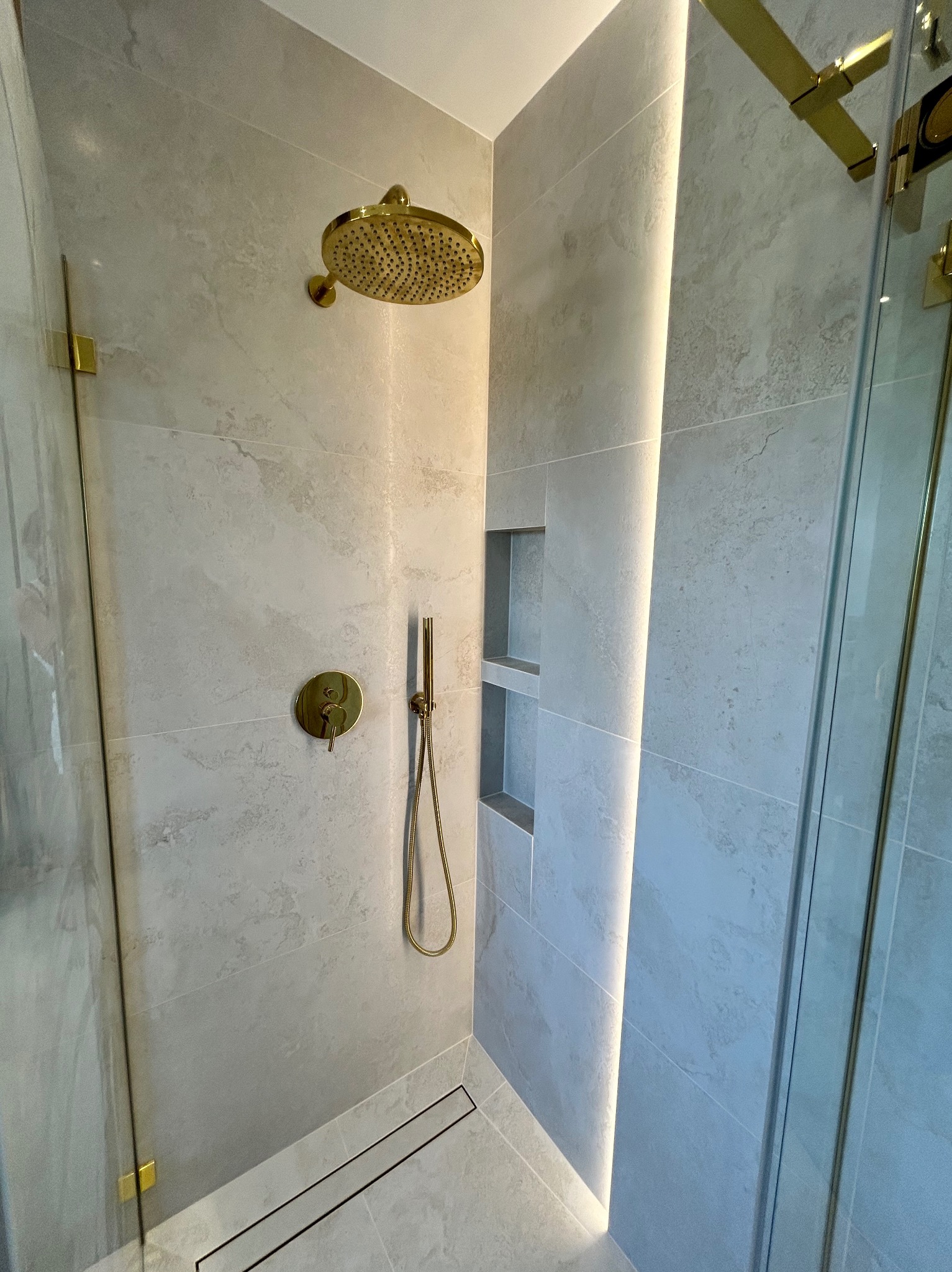 Nowoczesna kabina prysznicowa z elementami w kolorze złota, wykończona płytkami imitującymi marmur i oświetleniem LED, z odpływem liniowym.