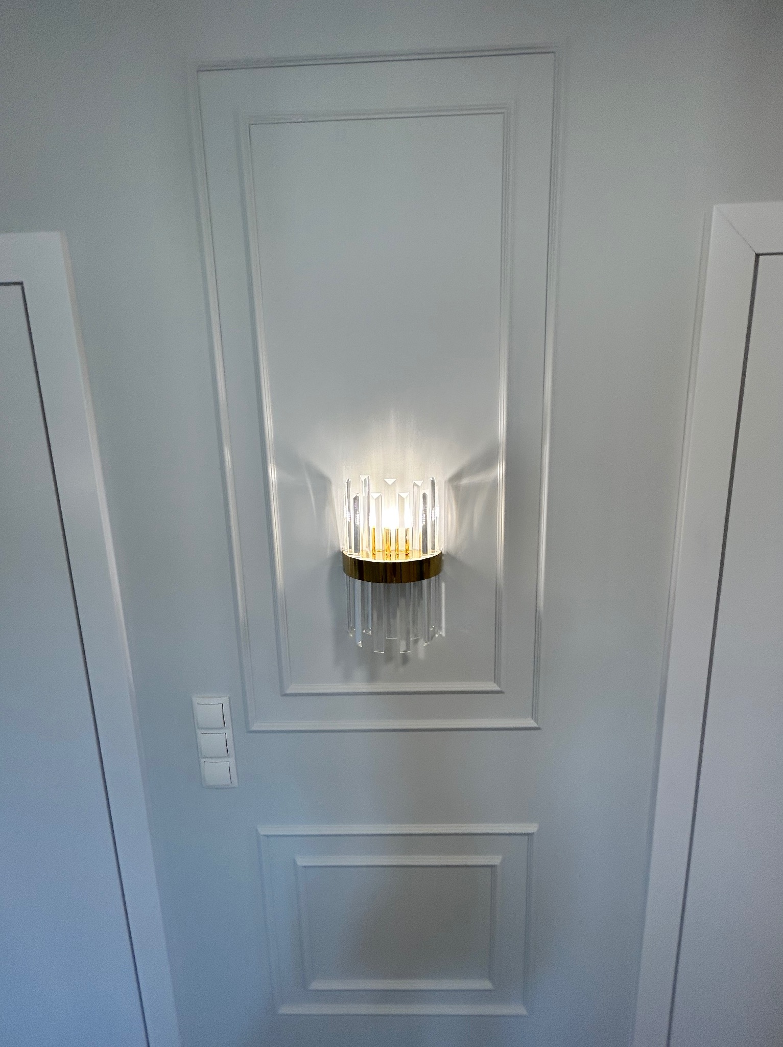 Biała ściana z dekoracyjną, podświetloną lampą ścienną z kryształkami i złotym elementem, obok widoczne drzwi i włącznik światła.