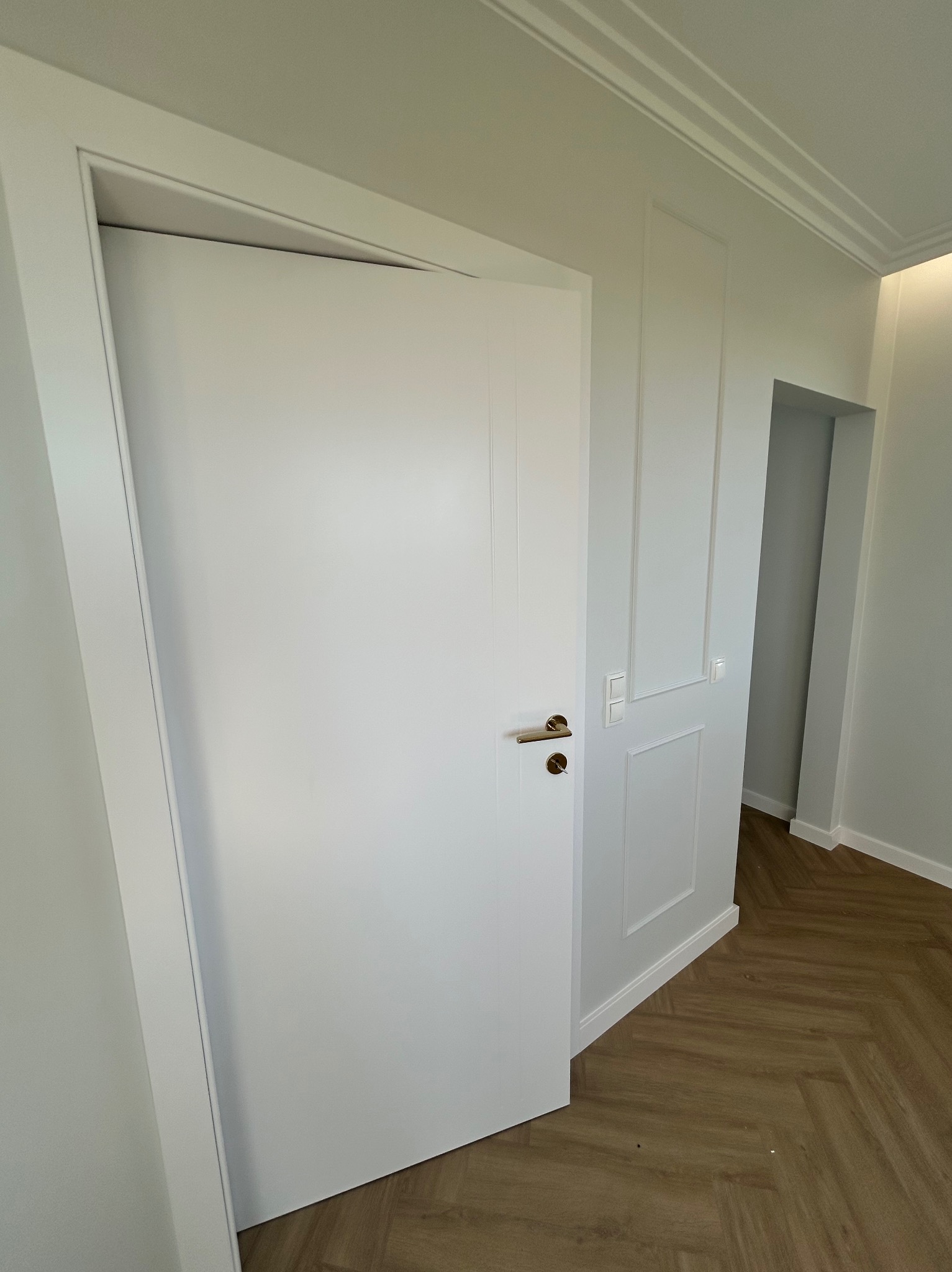 Białe drzwi wewnętrzne z złotą klamką, częściowo otwarte, na tle ściany z dekoracyjnymi panelami i parkietu w jodełkę, widok na korytarz.