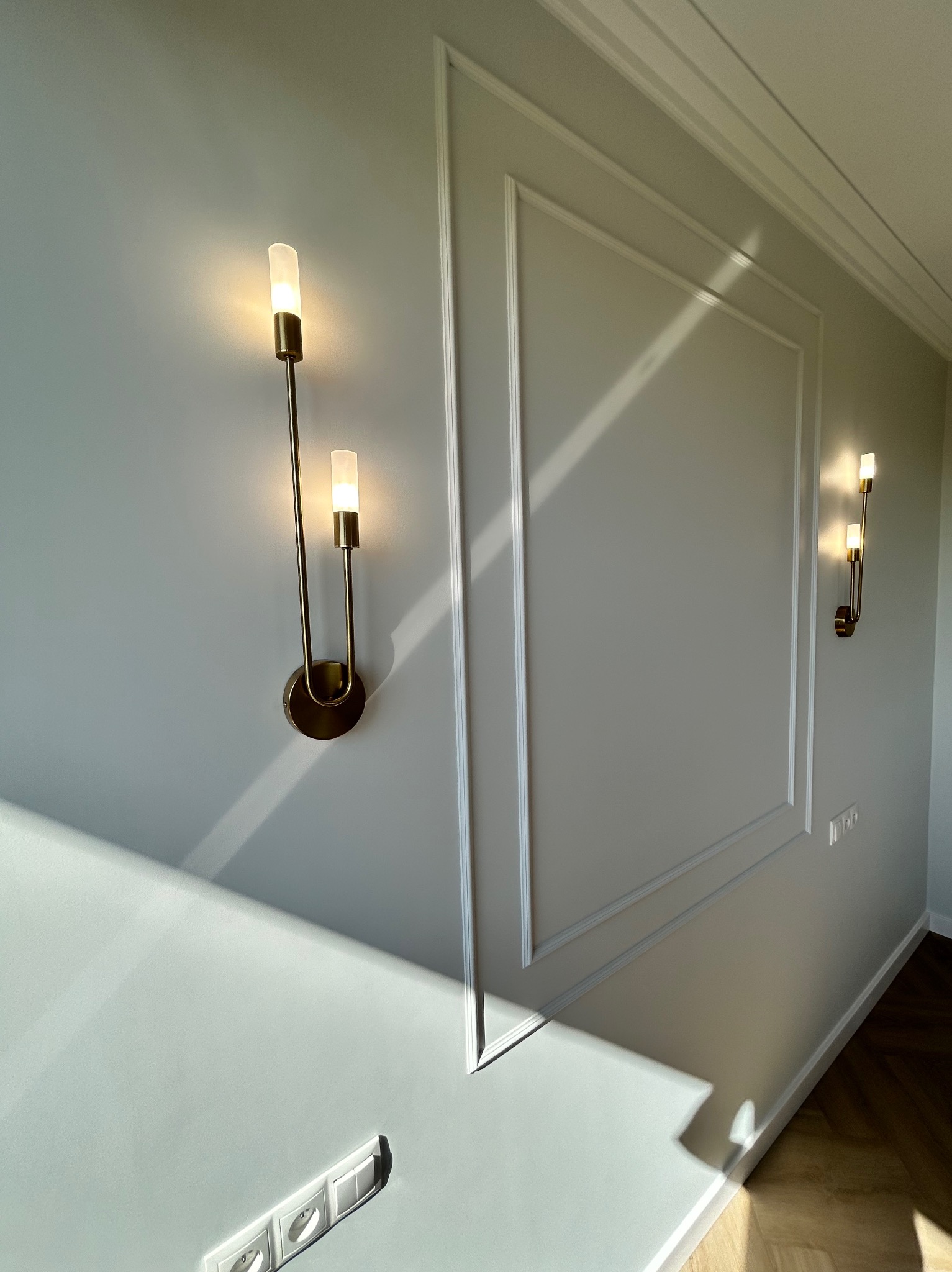 Ściana w jasnym odcieniu szarości z dekoracyjnymi listwami i dwoma złotymi kinkietami, oświetlona naturalnym światłem. Widoczne gniazdka elektryczne.