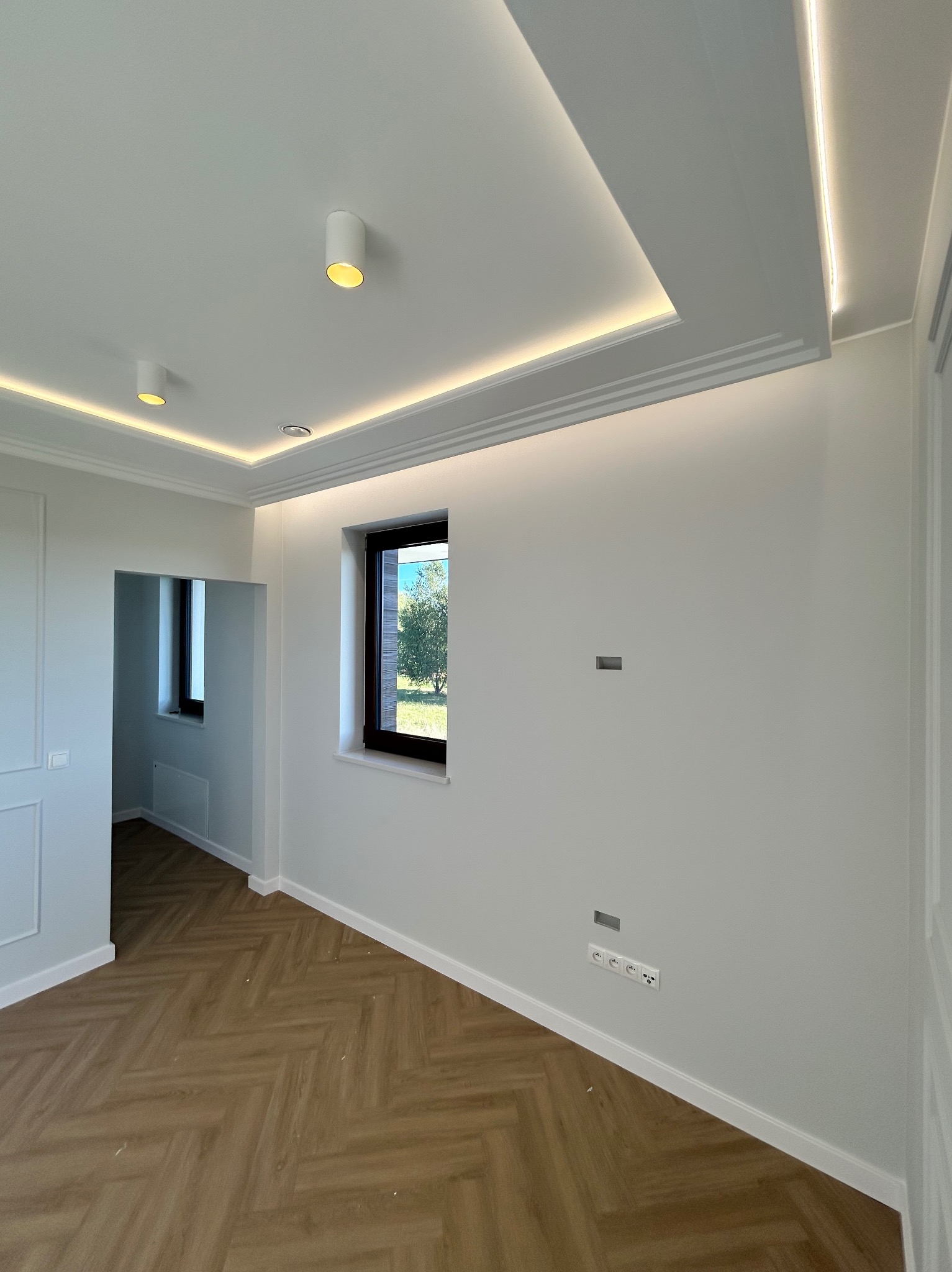 Jasne wnętrze po remoncie: parkiet w jodełkę, białe ściany z gniazdkami, okno z widokiem na zieleń i podwieszany sufit z oświetleniem LED.