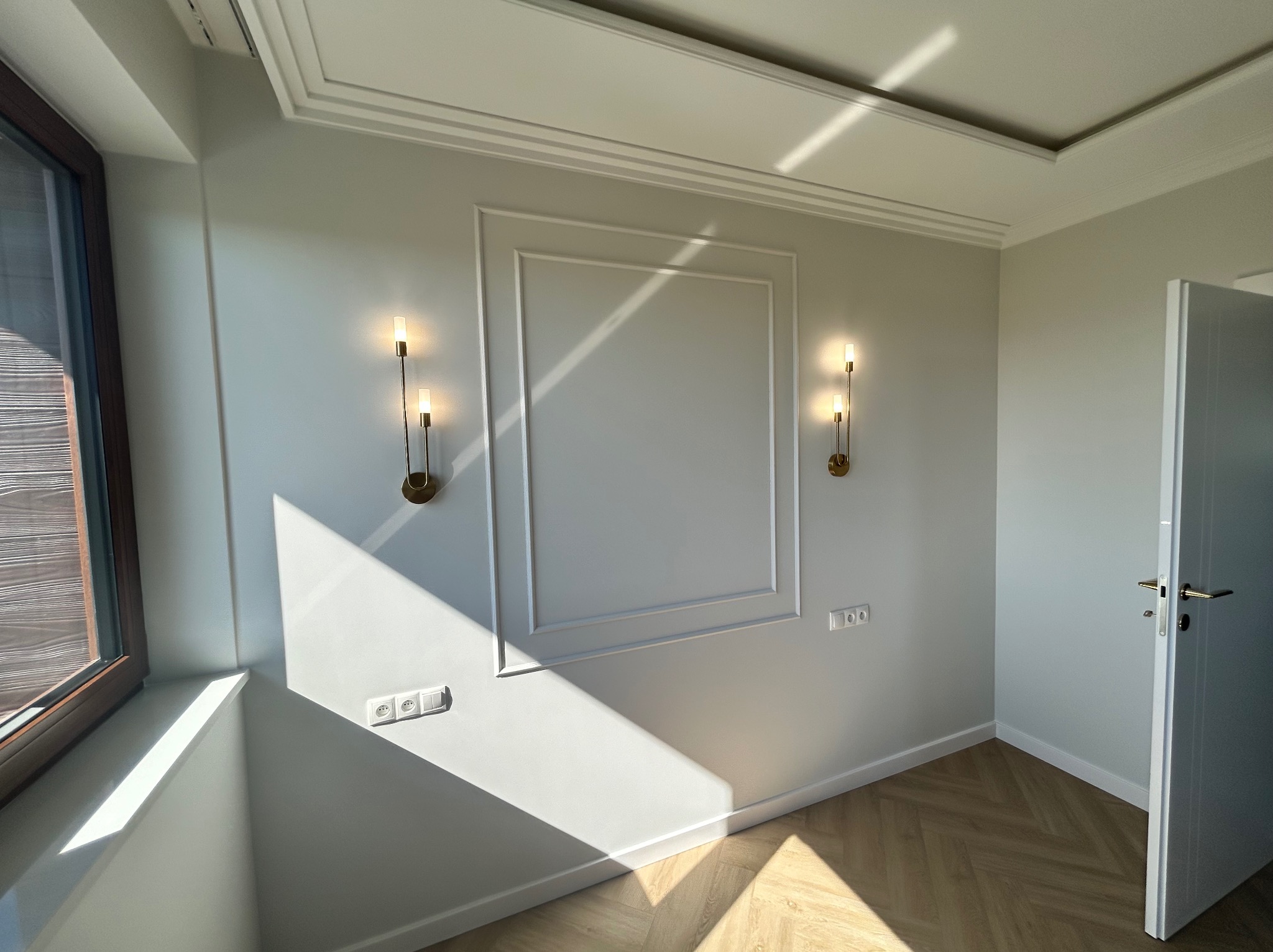 Jasne wnętrze: ściana z dekoracyjną ramą i kinkietami, parkiet w jodełkę, widok na okno i otwarte drzwi. Promienie słoneczne tworzą geometryczne cienie.