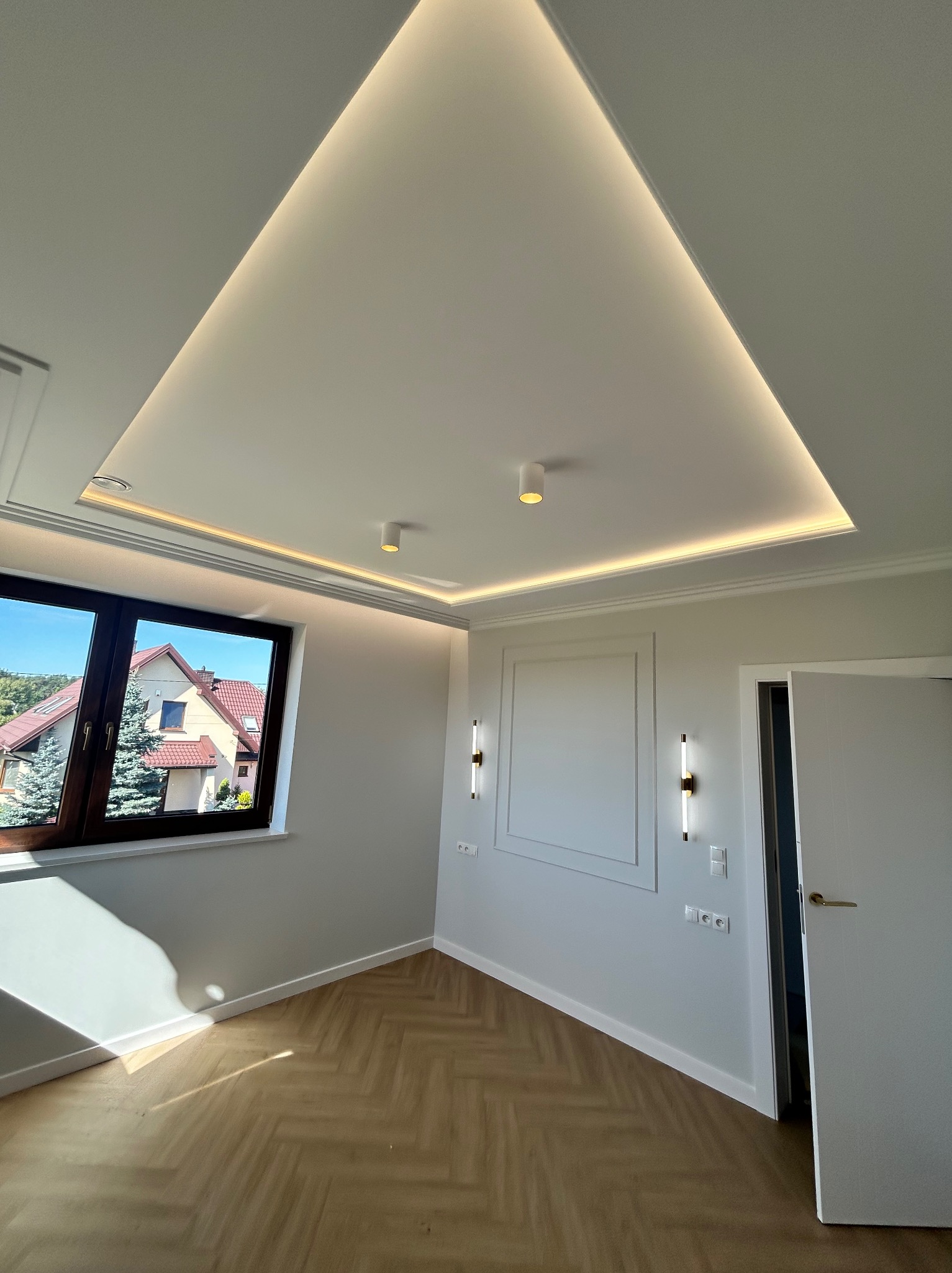 Nowoczesne wykończenie wnętrza: parkiet w jodełkę, białe ściany z dekoracyjną ramą, podwieszany sufit z oświetleniem LED. Widok na dom przez okno.