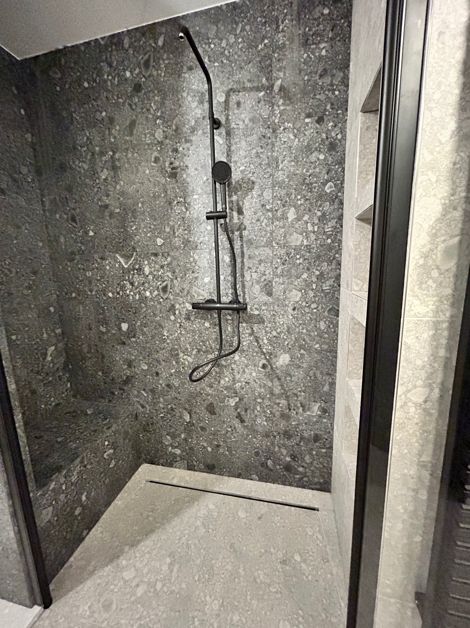 Nowoczesna kabina prysznicowa z czarną armaturą i szarymi płytkami imitującymi kamień, minimalistyczny design z odpływem liniowym.