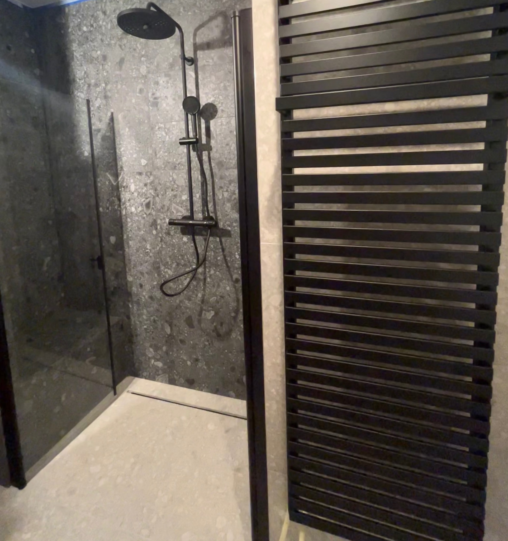 Nowoczesna łazienka z czarną armaturą: deszczownica, panel prysznicowy i grzejnik drabinkowy. Ściany wykończone płytkami imitującymi kamień.