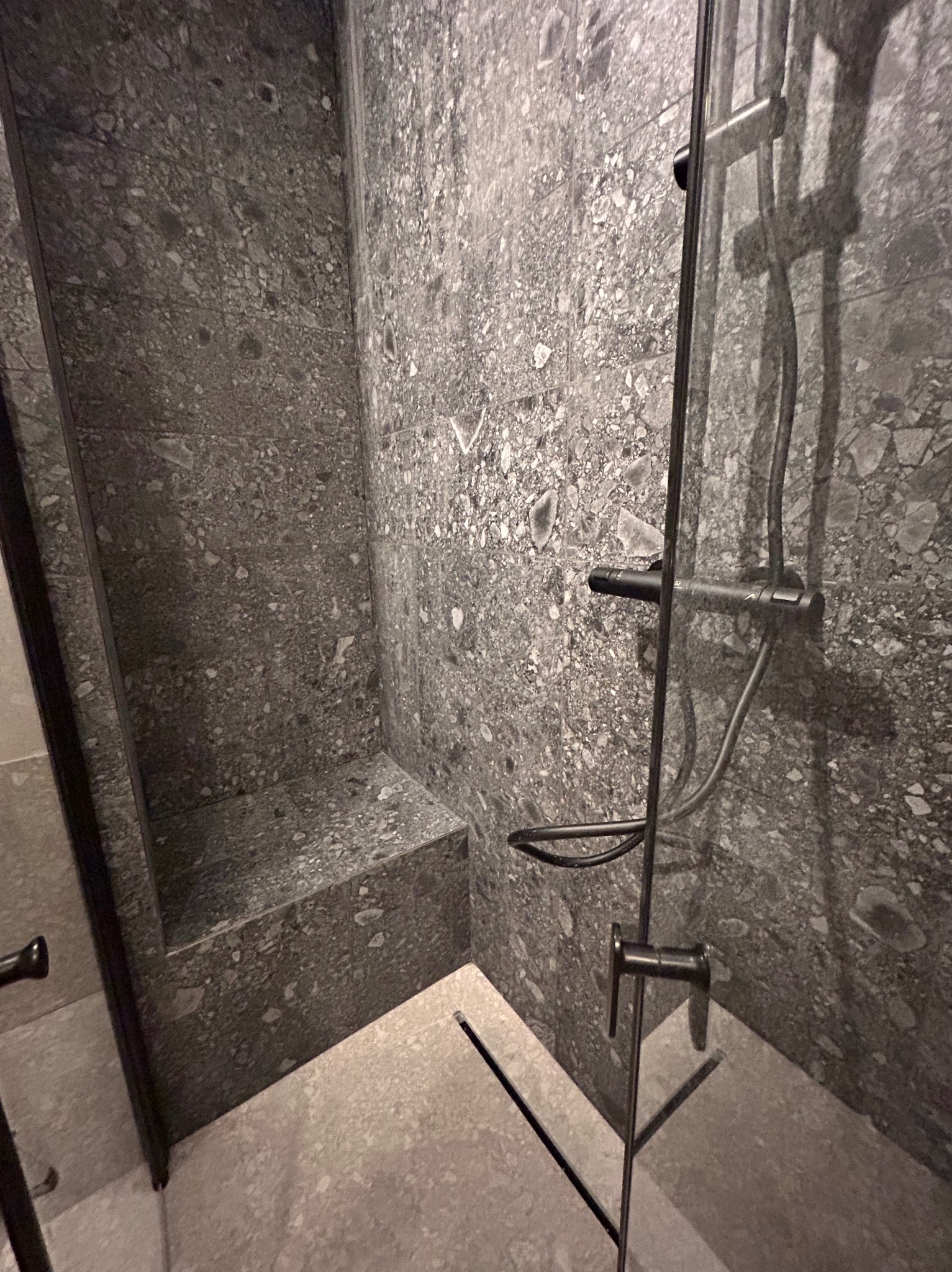 Nowoczesna kabina prysznicowa z szarymi płytkami imitującymi kamień, czarna armatura i odpływ liniowy. Minimalistyczny design z siedziskiem.