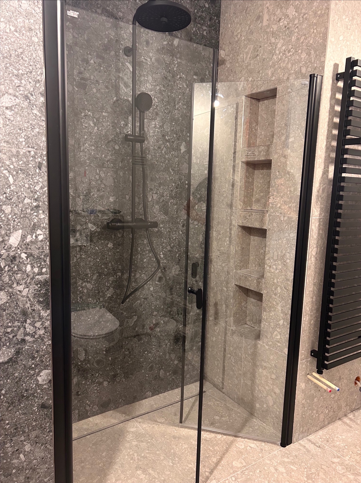 Nowoczesna kabina prysznicowa z czarnymi profilami i deszczownicą, wykończona płytkami imitującymi kamień. Widoczne wnęki w ścianie i grzejnik drabinkowy.