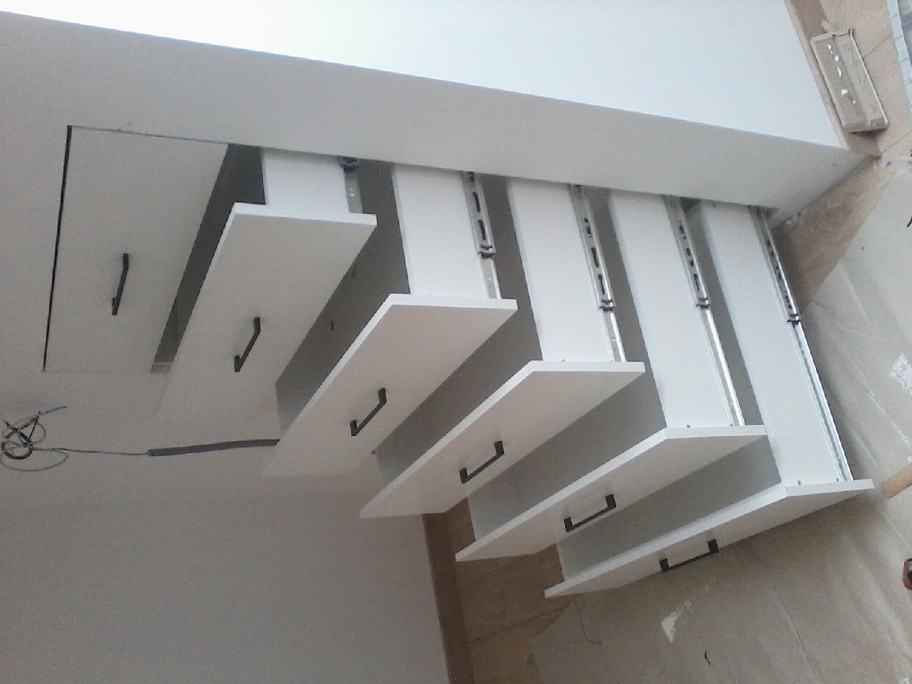 Białe, wysuwane półki z czarnymi uchwytami zamontowane pod sufitem, widoczne prowadnice i mocowania, wnętrze szafy wnękowej w trakcie montażu.