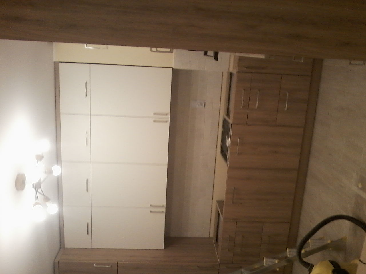 Nowoczesna zabudowa kuchenna z białymi frontami szafek górnych i drewnopodobnymi szafkami dolnymi, widoczny fragment blatu z płytą grzewczą, oświetlenie sufitowe w stylu industrialnym.