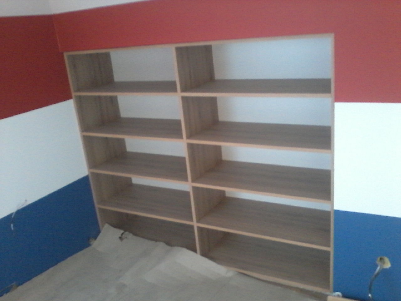 Pusta, drewniana szafa z półkami w trakcie montażu, zabezpieczona kartonem na podłodze, na tle ściany pomalowanej w poziome pasy: czerwony, biały i niebieski.