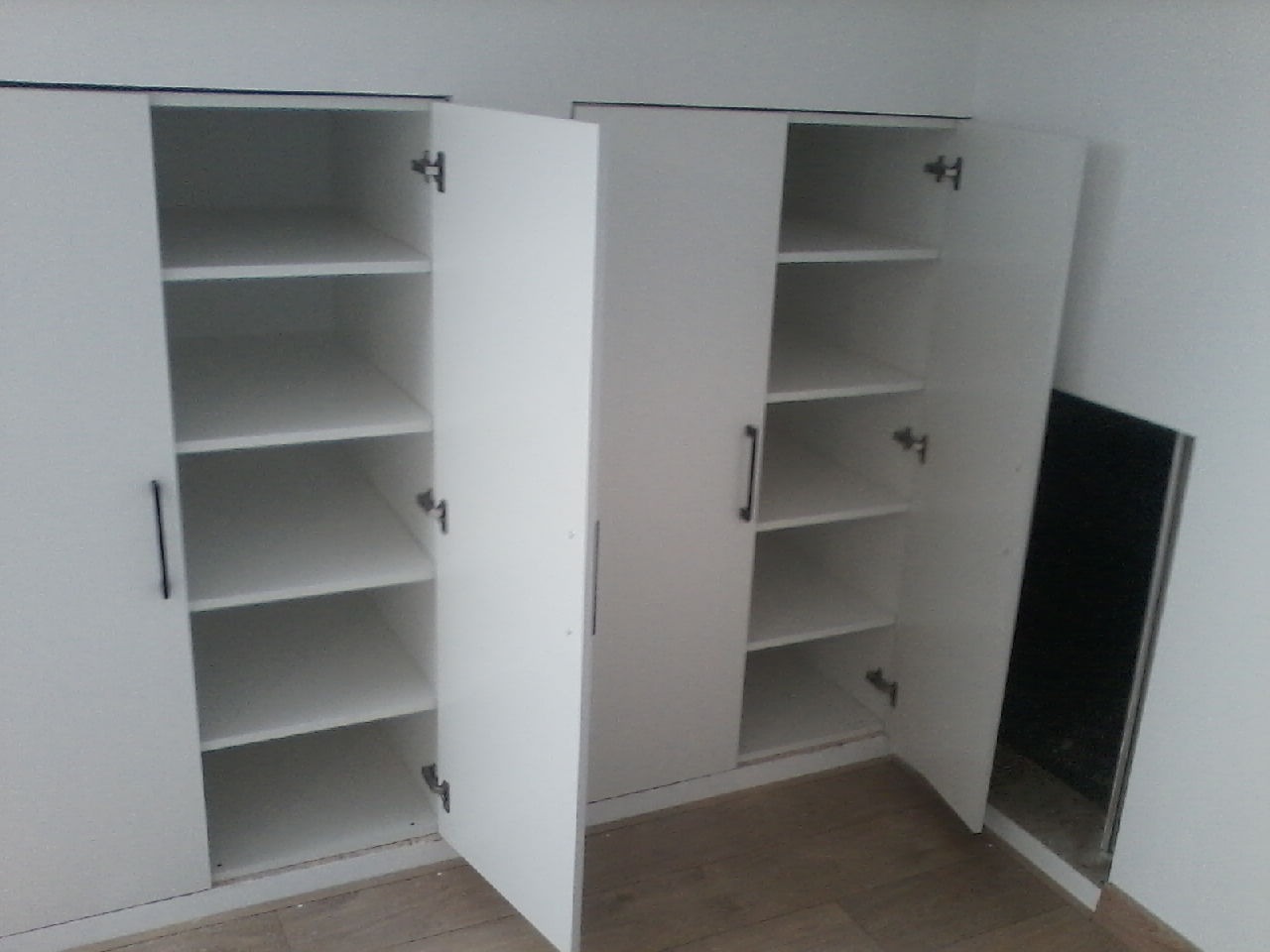 Biała szafa z otwartymi drzwiami, widoczne półki wewnątrz, minimalistyczny design, czarne uchwyty, podłoga imitująca drewno, fragment otworu wentylacyjnego w ścianie.