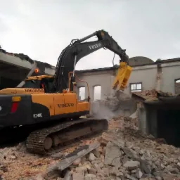 Żółto-czarna koparka Volvo EC240CL z hydraulicznymi nożycami do betonu podczas rozbiórki budynku z cegły, widoczne gruzy i fragmenty ścian.