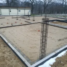 Przygotowany teren pod budowę domu z widocznymi fundamentami, szalunkiem i wystającymi zbrojeniami pionowymi, na obrzeżach leży śnieg.