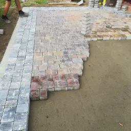 Układanie kostki brukowej z szarej i czerwonej kostki, widoczny fragment ułożonej nawierzchni i przygotowane podłoże, w tle fragment domu i stosy kostki.