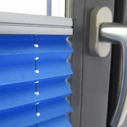 Niebieska plisa okienna zamontowana w szarej ramie okiennej z widoczną srebrną klamką.