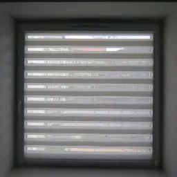 Okno z zamontowaną, częściowo opuszczoną, biało-szarą roletą dzień-noc z widocznymi poziomymi pasami, przez które widać fragment miejskiego krajobrazu za oknem.