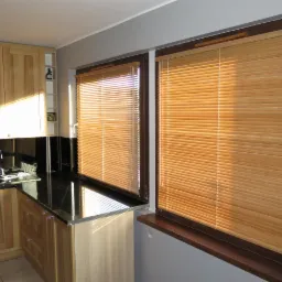 Jasne drewniane żaluzje poziome w oknach kuchni z drewnianymi szafkami i czarnym blatem, widok z wnętrza pomieszczenia.