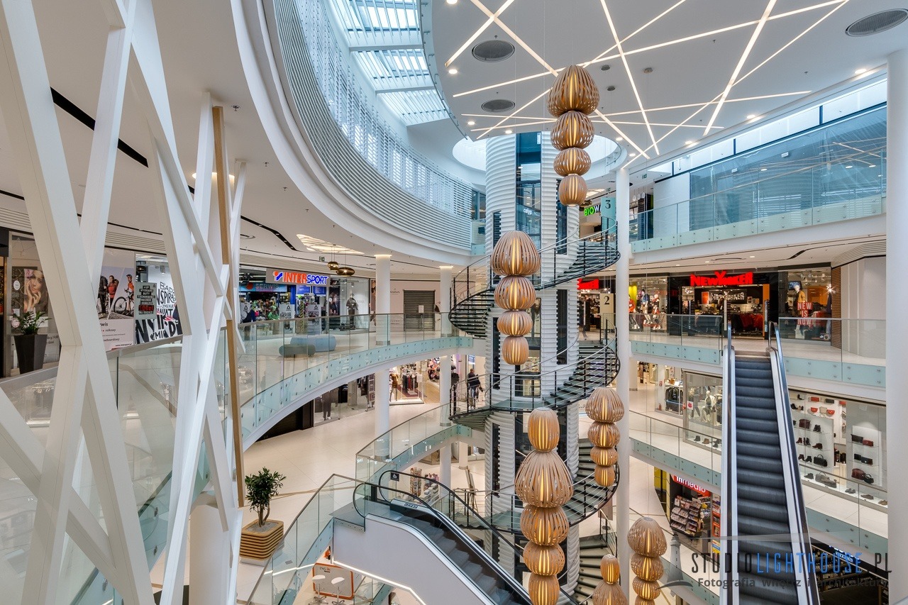 Wnętrze nowoczesnego centrum handlowego z wielopoziomową przestrzenią, spiralnymi schodami, ruchomymi schodami i dekoracyjnymi lampami wiszącymi.