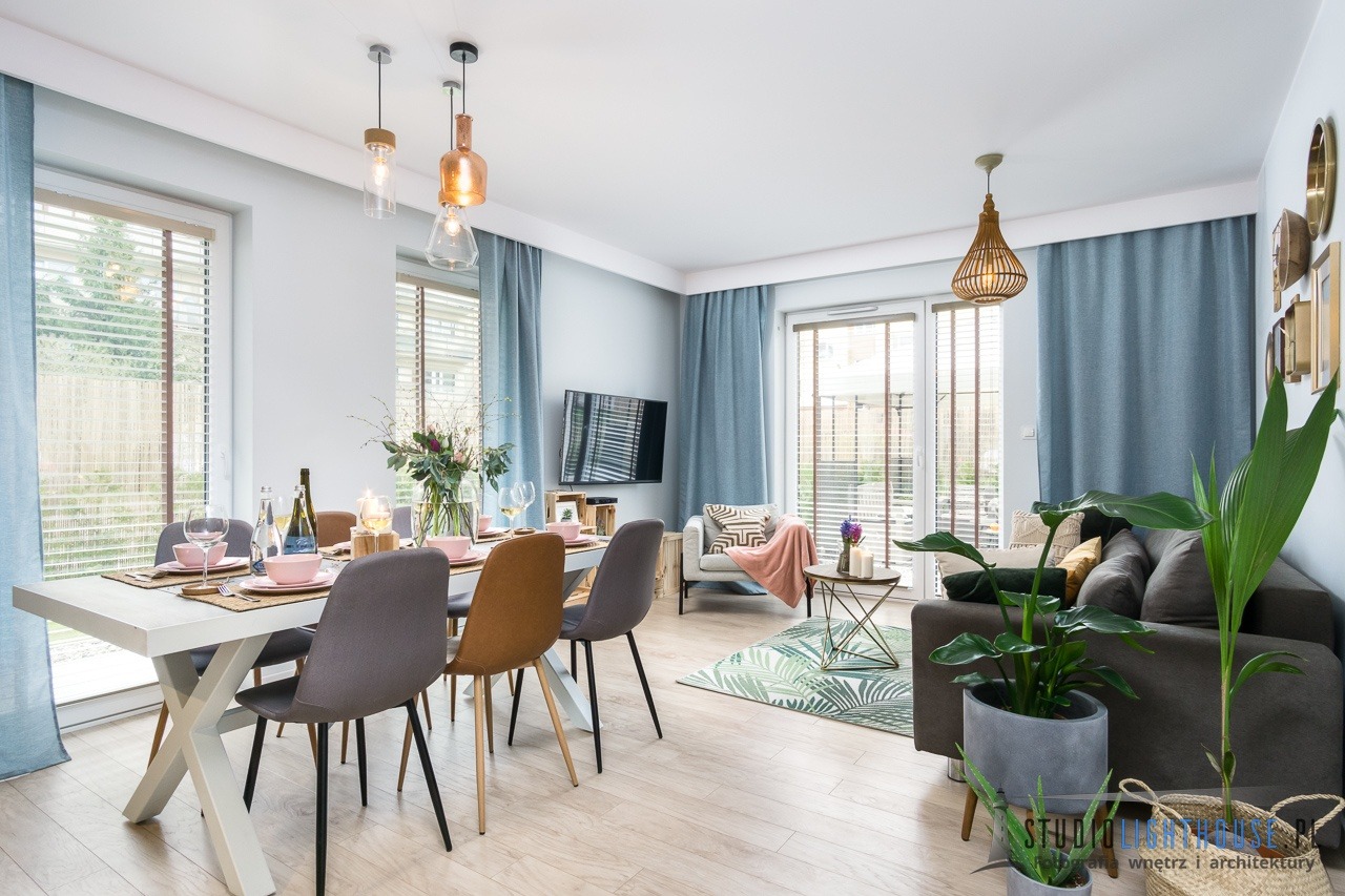 Jasne wnętrze salonu z jadalnią, stół z dekoracjami, szare krzesła, kanapa z roślinami w donicach, błękitne zasłony i drewniane żaluzje.